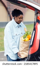 Vertical Portrait Of Young Black Woman Putting Grocery Bag In Car Trunk At Supermarket Parking Lot