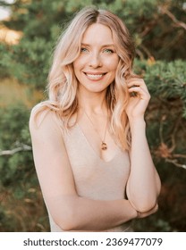 Vertical portrait of an young blonde happy smiling woman in beige dress posing in the woods near of pine trees. Foto Stok