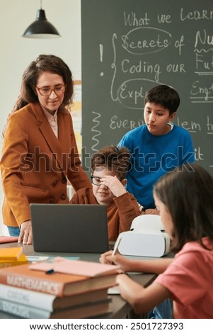 Vertical portrait of young boy using laptop in school classroom and setting up VR technology with female teacher helping