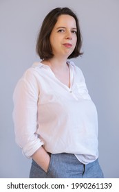 Vertical Portrait Of A Young Brunette Woman In White Shirt And Gray Trousers On Gray Wall Background. Business Portrait Of Employee, Manager, Casual Business Dress. Photo Presentation Of Employee.