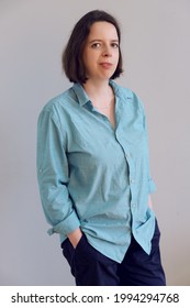 Vertical Portrait Of Young Brunette Woman In Turquoise Shirt And Blue Trousers On Gray Wall Background. Business Portrait Of Employee, Manager, Casual Business Dress. Photo Presentation Of Employee.