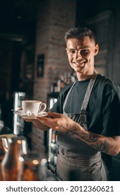 Vertical portrait of young smiling caucasian male barista serving hot aromatic coffee, cappuchino of latte in a white cup with marshmallow enjoying his job.