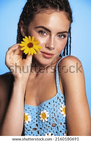 Vertical portrait of young tropical woman with dreads, crochet summer dreads, holding flower near glowing, moisturized clear facial skin, standing over blue background.