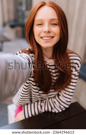 Vertical POV shot of happy young woman talking video call or taking selfie picture looking at camera sitting at table in outdoor cafe. Point of view of happy redhead female having video chat.