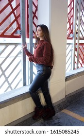 Vertical Profile View Of Pretty Brunette Woman With Waist Length Hair Wearing Black Leather Pants And Brown Sweater Looking Wistfully Out Of The Window Of An Indoor Parking Building