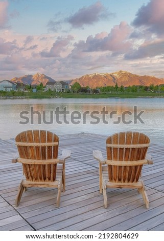 Vertical Puffy clouds at sunset Four wooden lounge chairs facing the reflective Oquirrh Lake at Daybreak, Utah. Wooden dock with a view of the residential area against the mountain range background.