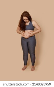 Vertical puzzled red haired overweight fat barefoot woman looking on tummy, doing activity, holding touching belly. Clothing plus size, skin folds. Turgor flabby skin lost. Beige isolated, copy space