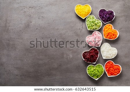 Vertical row of colorful heart-shaped bowls of grated vegetables and lots of grey metal surface for copy space to the side, studio shot made from above