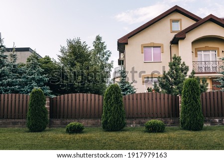 Vertical sections of brown metal profile fence. Live plantings. Green thuja, bushes and lawn. The local area decor. A house and a garden. Landscaping of the territory. Capital fencing. Vacation home.