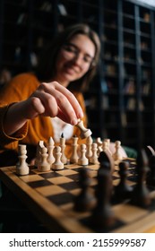Vertical selective focus shot of smiling woman wearing elegant eyeglasses making chess move sitting on armchair in dark library room. Pretty intelligent lady playing logical board game alone at home.