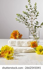 Vertical setup with stacked marble blocks, marigold, yellow flowers, and green branches. Natural light highlights the organic wellness concept and space for product placement.