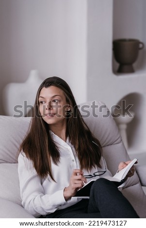 Vertical shoot of successful designer, pretty woman sitting on cozy chair at home looking aside, dressed in white shirt and black pants holding drawing pad. Attractive brunette woman relaxing at home.
