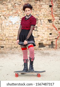 Vertical Shot Alternative Emo Girl Sitting On A Skate Board On A Brick Wall Background. Dark Teen In A Black Skirt, Striped Stocking And Steel Boots