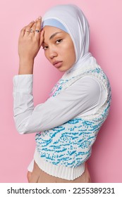 Vertical shot of attractive woman keeps hands on forehead looks at camera with serious expression wears headscarf white shirt and vest rings on fingers keeps to certain religion isolated on pink wall - Shutterstock ID 2255389231
