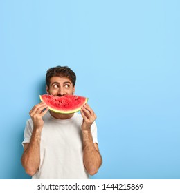 Vertical Shot Of Attractive Young Man Covers Mouth With Slice Of Red Juicy Watermelon, Enjoys Eating Seasonal Tropical Fruit During Summer Period, Wears White T Shirt, Poses Indoor. What Nice Taste!