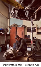 Vertical Shot Of Auto Mechanic Squatting Working On Car Detail On Floor At Auto Repair Shop Under Lifted Vehicle. Repairman In Dirty Workwear. Tools, Spare Parts And Garage Interior On Background