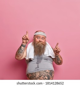 Vertical Shot Of Bearded Caucasian Adult Man Gets Sweaty During His Training Session, Works Hard On Losing Excess Weight And Points Above. Funny Obese Guy Wears Headband Shirt, Goes In For Sport