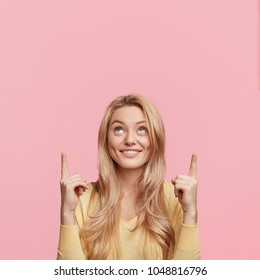 Vertical shot of beautiful female with attractive look indicates happily upwards with forefingers, points at balnk copy space for your advertisment or promotional text. Just look there, please!