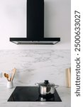 Vertical shot with black extractor hood over modern glass ceramic cooker with stainless steel pan. Saucepan on electric induction stove in kitchen interior. Marble tile, white copy space wall