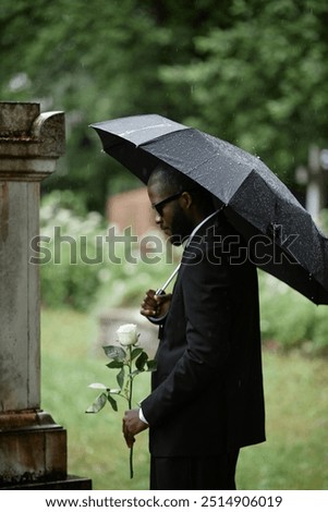 Vertical shot of Black man grieving over loss of relative at cemetery next to grave holding white rose to commemorate deceased and hiding from rain under black umbrella, copy space