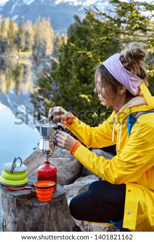 Vertical shot of busy female hiker makes aromatic coffee on camping stove, boils water, poses near stump with teacup, has vacation in mountains near beautiful lake. Tourism equipment and tools concept