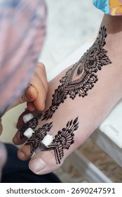  A vertical shot capturing the process of a beautiful, ornate black henna design being drawn on the skin of a foot and toes. The image highlights traditional body art and the artist's  focused work.