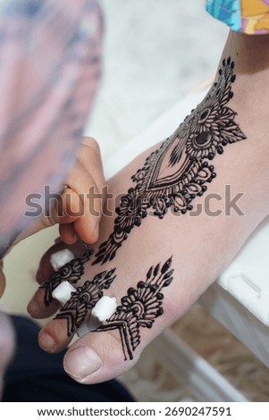  A vertical shot capturing the process of a beautiful, ornate black henna design being drawn on the skin of a foot and toes. The image highlights traditional body art and the artist's  focused work.