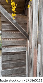 A vertical shot capturing the unique architectural details of a rustic building made from aged wooden planks. The exterior walls feature a mix of horizontal and vertical timber siding with natural textures and weathered grey brown tones. A prominent diagonal wooden beam adds dynamic geometric lines to the composition. Bright yellow flowers drape gracefully from the roof area providing a beautiful natural contrast against the rugged timber surfaces. This high quality photograph showcases traditional craftsmanship and organic materials in a modern garden setting. The interplay of light and shadow on the various wood grains creates a rich visual depth ideal for architectural blogs interior design concepts or nature themed backgrounds. Each plank shows signs of age and history giving the structure a soulful and authentic character.