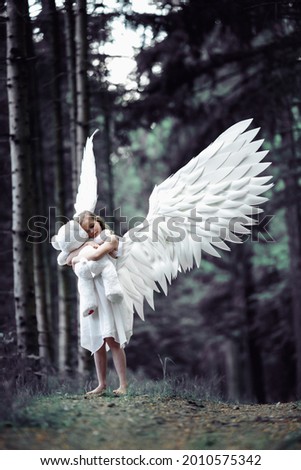 A vertical shot of a Caucasian blonde girl in angel wings costume with a teddy bear toy