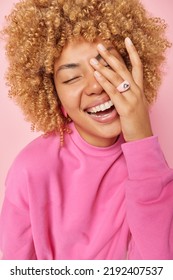Vertical shot of cheerful carefree woman has curly blonde hair makes face palm smiles broadly keeps eyes closed expresses positive feelings wears pink pullover poses indoor. People emotions concept - Shutterstock ID 2192407537