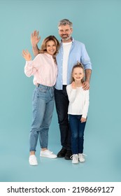 Vertical Shot Of Cheerful Family Waving Hands To Camera Standing Over Blue Studio Background. Middle Aged Parents And Kid Daughter Gesturing Hello. Full Length