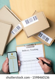 Vertical shot. Concept of order tracking system. Delivery man holding smartphone and writing down some details about order, cardboard boxes on the table