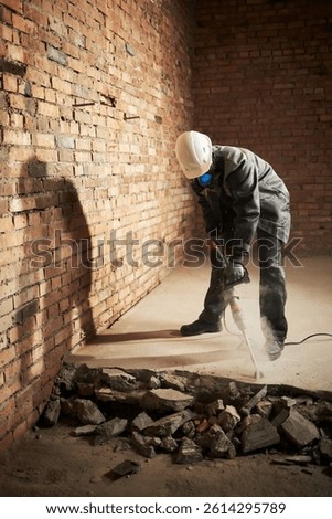 Vertical shot of construction worker using new jackhammer to destroy cement on floor, he wearing uniform and respirator to protect himself from dust