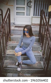 A Vertical Shot Of A Cool, Fashionable Teenager With Blue Sunglasses Sitting On An Apartment Front Steps