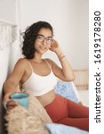 Vertical shot cozy young gorgeous easter woman resting on rattan couch with cute pillows, wear comfortable yoga leggings, sportsbra, holding mug, enjoying fresh coffee smiling, relaxing weekends