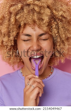 Vertical shot of curly haired feamle model uses tongue scraper takes care of oral hygiene dressed in purple t shirt cleans mouth poses indoors. Young beautiful woman uses tongue cleaner regularly