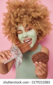 Vertical Shot Of Curly Haired Woman Winks Eye Applies Nourishing Clay Mask To Unclog Pores Bites Sweet Bar Of Chocolate Holds Delicious Cookie Enjoys Eating Sugary Food Isolated Over Pink Wall