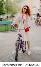 Vertical shot of curly red haired woman rides bicycle in city during weeked, has free time, wears long dress, shades and sneakers, carries small red bag, keeps hands on gear. Recreation concept