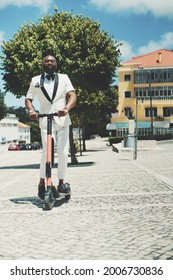 A vertical shot of a dapper adult bearded bald black senior in eyeglasses and a white fancy summer costume is holding in hands the handlebars of the e-scooter ready to ride over the paving-stone