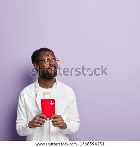 Vertical shot of dark skinned guy passenger holds passport with fly ticket, going to have business trip, wears spectacles and white shirt, focused above, poses against purple wall, blank space upwards