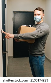 Vertical Shot Of Delivery Man In Medical Mask Ringing Doorbell Of Customer Apartment Looking At Camera, Holding Cardboard Box And Contactless Payment POS Wireless Terminal For Card Paying.