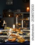 Vertical shot of dining table set with variety of festive dishes, champagne glasses and lit candles while christmas lights are glowing in background, copy space