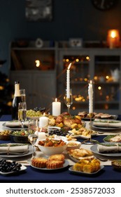Vertical shot of dining table set with variety of festive dishes, champagne glasses and lit candles while christmas lights are glowing in background, copy space