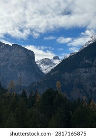 A vertical shot of a dramatic mountain valley framed by steep, dark rock cliffs and dense evergreen forest in the foreground. In the center distance, snow-capped alpine peaks rise under a partly cloudy, bright blue sky.