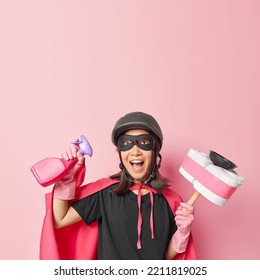 Vertical Shot Of Emotional Superwoman Exclaims Loudly Holds Cleaning Detergent Plunger With Toilet Paper Brings House In Order Dressed In Superhero Costume Isolated Over Pink Studio Background