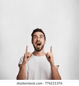 Vertical shot of excited surprised male has beard and mustache indicates with fore fingers upwards, advertises something amazing, stands against white concrere wall with copy space for promotion