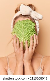 Vertical shot of faceless woman covers face with fresh green cabbage leaf rejuvenates skin uses vegetable wears soft headband stands bare shoulders against brown background. Natural beauty concept