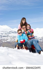 Vertical Shot Of A Family Of Four Sitting On A Snow Sled In A Snowy Field On A Bright, Sunny Day And Embracing Each Other