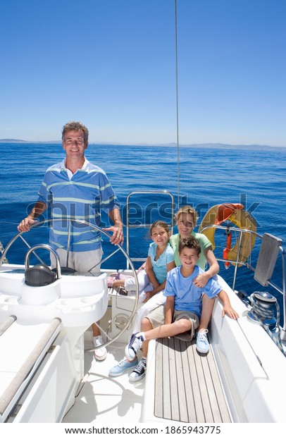 Vertical shot of a\
family relaxing on the deck of a sailing boat out at sea with the\
father standing at the\
helm.