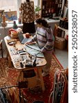 Vertical shot of female sales assistant folding clothes on wooden display table, cluttered with vintage fashion accessories at second hand shop with retro style carpet on floor